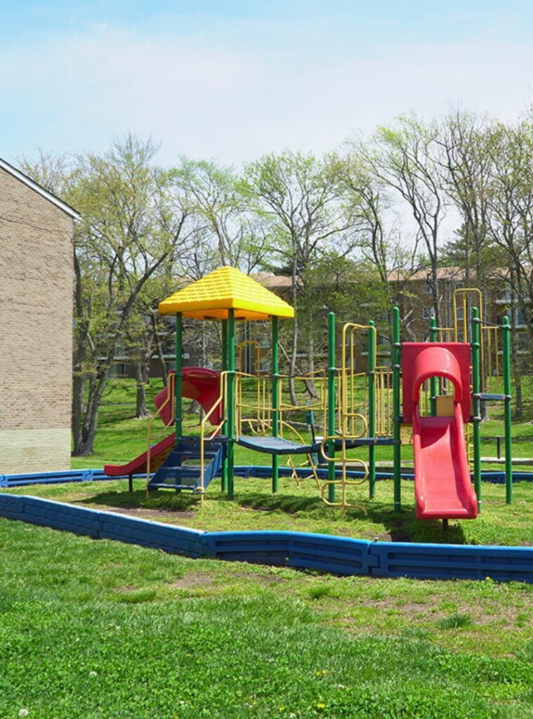 A colorful playground with a red slide, yellow canopy, and green bars, surrounded by grass and a building on the left.